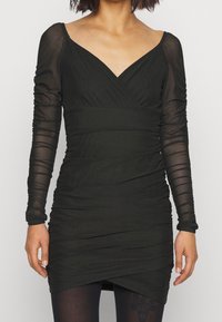 Kvinna iklädd en rynkad svart miniklänning med genomskinliga långa ärmar och V-ringning, tillsammans med mörka mönstrade strumpbyxor mot en enfärgad bakgrund.