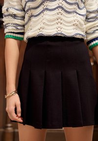 Jupe plissée noire en tissu doux, présentant une texture lisse et des lignes épurées. Associée à un pull texturé de couleur claire.