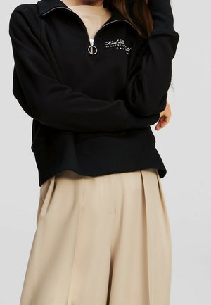Personne portant un sweat-shirt noir à fermeture éclair quart de zip avec texte blanc et pantalon beige plissé à jambes larges sur un fond uni.