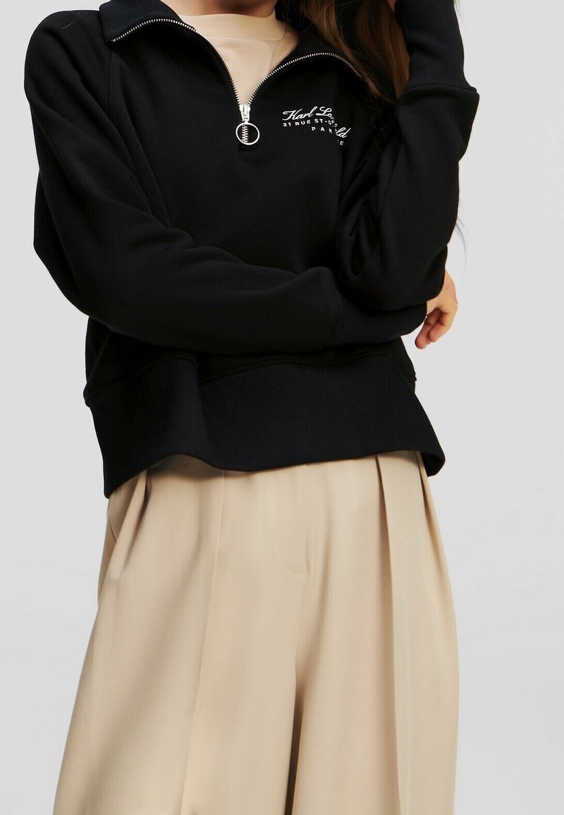 Personne portant un sweat-shirt noir à fermeture éclair quart de zip avec texte blanc et pantalon beige plissé à jambes larges sur un fond uni.