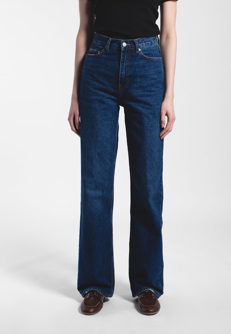 Person trägt hoch taillierte, dunkelblaue Jeans mit geradem Bein, ein schwarzes kurzärmeliges Oberteil und braune Lederschuhe vor einem einfarbigen Hintergrund.