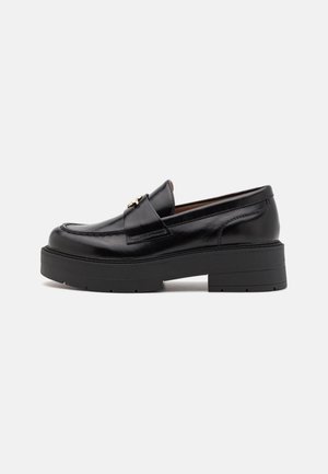 Mocassin à plateforme en cuir noir avec semelle épaisse et détail en métal doré sur le dessus, vue de profil sur fond gris clair.