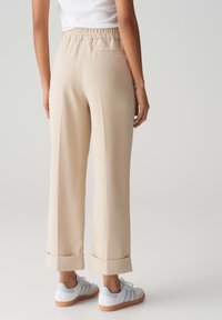 Pantaloni beige a gamba larga con cintura elastica, una tasca posteriore singola e orli arrotolati, abbinati a sneakers bianche su uno sfondo chiaro.