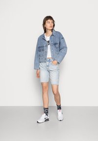 Chaqueta de mezclilla en azul claro con dos bolsillos frontales, combinada con pantalones cortos de mezclilla azul claro y zapatillas blancas. Textura suave, diseño informal.