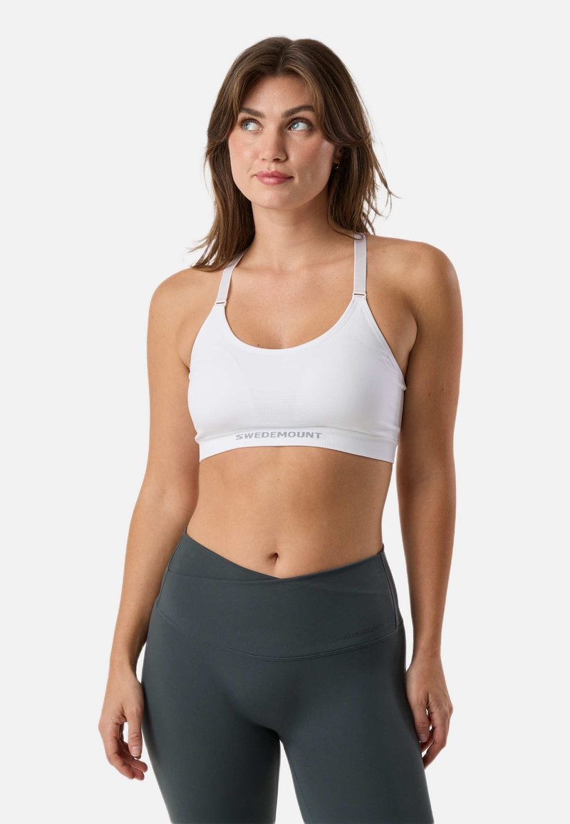 Vrouw in een witte sportbeha en donkergrijze legging kijkt omhoog en iets opzij tegen een effen achtergrond.