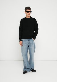Zwarte gebreide trui met een ronde hals, gecombineerd met lichtblauwe wijdvallende jeans. Model draagt ook zwarte zonnebril en zwarte schoenen.