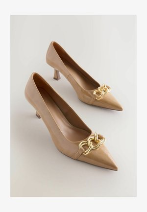 Scarpe con tacchi alti in pelle verniciata beige, con punta affusolata e dettagli decorativi in catena dorata sul davanti, su uno sfondo chiaro.