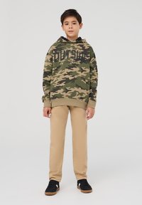 Bluza kamuflażowa w zielonym i beżowym kolorze z czarnym napisem, w połączeniu z beżowymi spodniami i czarnymi trampkami na gumowej podeszwie. Styl casualowy.