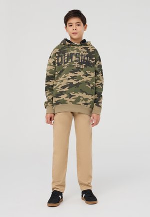 Felpa camouflage verde e beige con lettering nero, abbinata a pantaloni beige e sneakers nere con suola in gomma. Vestibilità e design casual.