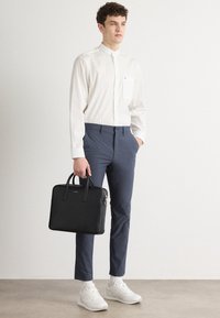 Una valigetta in pelle nera con manici superiori, indossata con una camicia bianca, pantaloni blu navy e sneakers bianche su uno sfondo neutro.