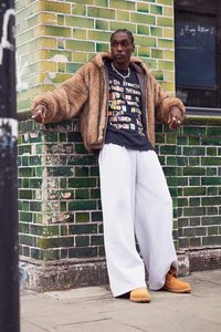 Person som lener mot en grønnflislagt vegg iført en brun pelskåpe, grafisk svart skjorte, vide hvite bukser og brune støvler.