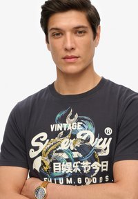 Mørkeblå t-shirt lavet af bomuld, med et grafisk design af koi-agtige fisk og bølger i gul og blå, med hvid tekst og rund halsudskæring.
