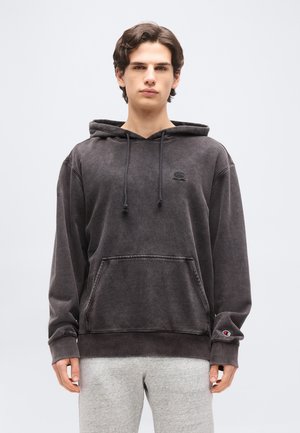 Fiatal férfi hullámos barna hajjal, kopott fekete kapucnis pulóvert visel elöl zsebfedéllel és világosszürke melegítőnadrágot, egy sima háttér előtt állva.