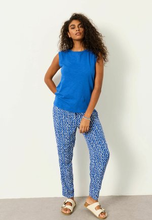 Kvinde med krøllet hår iført en ærmeløs blå top, blå bukser med hvide bølgemønstre og beige sandaler, stående op ad en hvid væg.