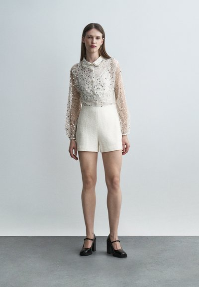 Femme portant un chemisier transparent à manches longues orné de perles, un short blanc taille haute et des talons noirs de style Mary Jane, debout devant un fond uni.