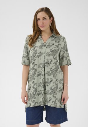 Nő világoszöld, paisley mintás, gombos ingben, rövid ujjúban és sötétkék térdig érő rövidnadrágban.