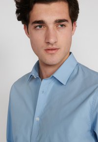Jaunas vyras su trumpais rudais plaukais, vilkintis šviesiai mėlyną marškinį su sagomis, žiūri šiek tiek į šoną prieš paprastą foną.