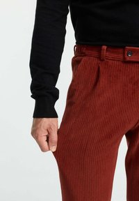 Rote Cordhosen mit vertikaler Rippung, ausgestattet mit einem Bund und einem Knopf. Die Hand greift den Stoff des Beins. Ein schwarzer Strickpullover ist sichtbar.