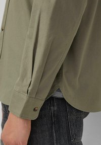 Een mouw in een olijfgroen, aangeknoopte blouse over donkere jeans, met een gestreept shirt dat aan de taille zichtbaar is.
