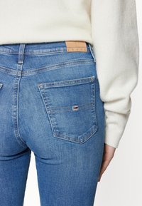 Blå denimjeans med medelhög midja, utrustad med två bakfickor och en tan läderlogotyp på midjebandet. Mjuk textur och smal silhuett.