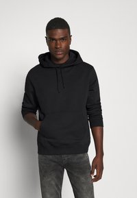 Svart hoodie med lång ärm, känguruficka och dragsko. Har en mjuk yta och avslappnad passform. Bärs med grå jeans.