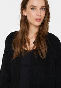 Svart strukturerad cardigan i mjukt tyg, med öppen framsidan, långa ärmar och en avslappnad passform. Bärs över en svart topp.