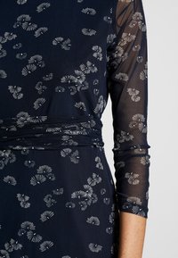 Marineblaues Kleid mit floralem Druckdesign; verfügt über transparente lange Ärmel und eine geraffte Taille. Strukturierter Stoff mit zarten weißen Blattmustern.