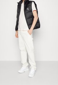 Svart vadderad väst med dragkedja, CK-logga på bröstet, kombinerad med en ljusrosa t-shirt, vita byxor och vita sneakers. Slät textur, avslappnad passform.
