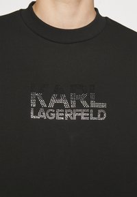 Felpa nera con colletto alto che presenta la scritta "KARL LAGERFELD" in luccicanti strass argento. Tessuto morbido.