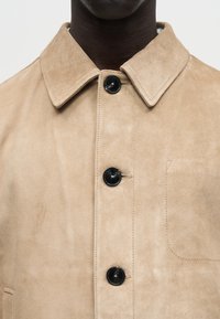 Κοντινό πλάνο σε ταμπα τζάκετ από σουέτ με μαύρα κουμπιά, φαρδύ γιακά και διακριτική τσέπη στο στήθος, που φοριέται από άτομο με σκούρο δέρμα.