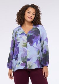 Blusa floreale di colore azzurro chiaro con grandi fiori viola e verdi, maniche lunghe, scollo a V e dettaglio della chiusura frontale con bottoni. Tessuto morbido e leggero.
