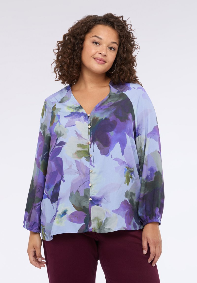 Blusa floreale di colore azzurro chiaro con grandi fiori viola e verdi, maniche lunghe, scollo a V e dettaglio della chiusura frontale con bottoni. Tessuto morbido e leggero.