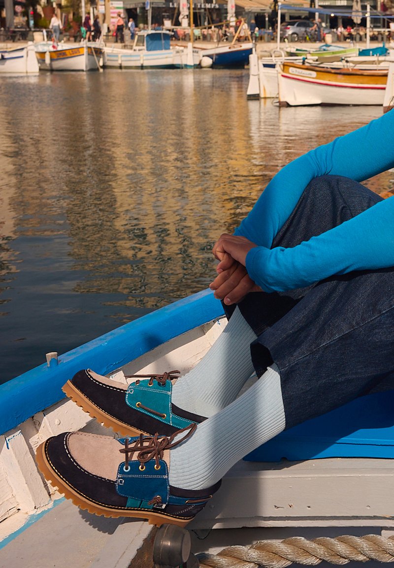 Personne portant des chaussures bateau bleues, beiges et noires avec des chaussettes blanches, assise sur un bateau à côté d'une marina avec des bateaux amarrés et de l'eau en arrière-plan.