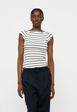 Femeie purtând un top alb ajustat cu mâneci scurte și dungi orizontale negre și pantaloni negri cu talie înaltă, stând în fața unui fundal simplu.