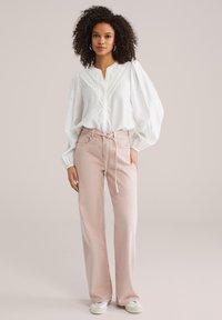 Blouse blanche texturée avec manches bouffantes et patte partielle, associée à un pantalon taille haute rose clair et des baskets blanches.