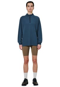 Długi rękaw, rozpinany pulower w ciemnoniebieskim teksturowanym materiale, w zestawieniu z beżowymi szortami rowerowymi i czarnymi butami sportowymi w białych skarpetach.