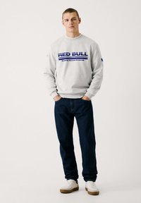 Sweat à capuche gris avec le logo "RED BULL" en bleu, jeans bleu marine, et baskets blanches avec des accents beige. Le mannequin se tient les mains dans les poches.