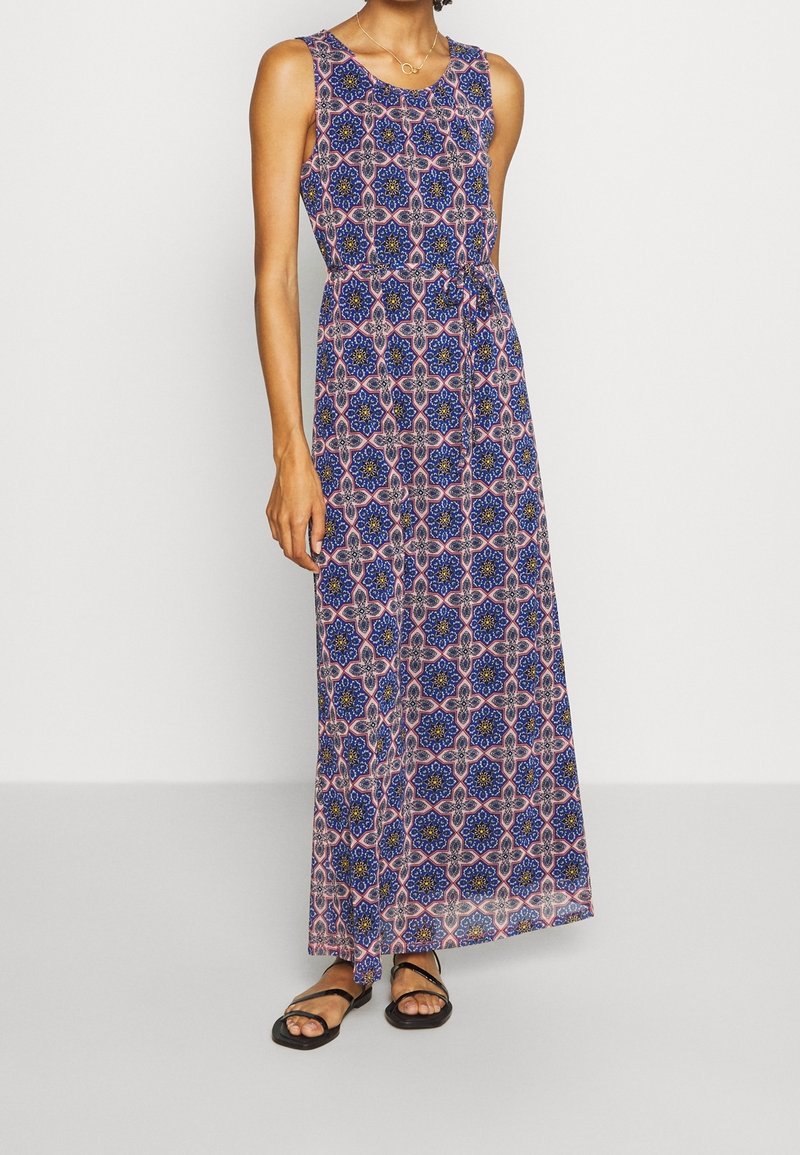 Femme portant une robe longue sans manches à motif floral géométrique bleu et rose, avec des sandales noires à brides.