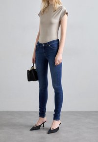 Beige aansluitend topje, donkerblauwe skinny jeans, zwarte puntige hakken met een kleine hak en een klein zwart handtasje. Glad materiaal, modern ontwerp.
