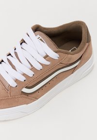 Zapatillas de ante marrón claro con cordones blancos, una lengüeta de tela texturizada y una franja de acento blanca y marrón oscuro a lo largo del lado.
