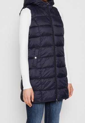 Nainen, joka pukeutuu tummansiniseen toppattuun hihattomaan liiviin, jossa on etuvetoketju ja painonapitaskut, valkoinen pitkähihaiseen paitaan ja sinisiin farkkuihin, seisoo valkoista seinää vasten.