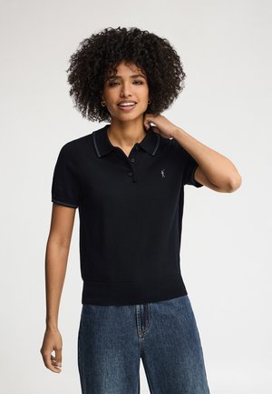 Frau mit lockigem Haar, die ein schwarzes Poloshirt mit kurzen Ärmeln und blaue Jeans trägt, lächelt und berührt mit einer Hand ihren Hals.