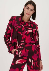 Bloemenpak in donkerburgundy met gedurfde roze en witte patronen. Beschikt over een getailleerde blazer met een reverskraag en bijpassende broek.