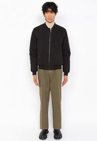 Schwarze Bomberjacke mit Frontreißverschluss, gerippten Bündchen und Saum, kombiniert mit olivgrünen Hosen mit geradem Bein und schwarzen Schuhen.