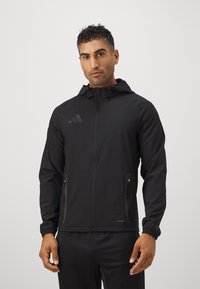 Zwarte sportjack met capuchon. Voorzien van ritszakken, elastische manchetten en een gladde, lichte stof. Subtiele logodetail op de borst.