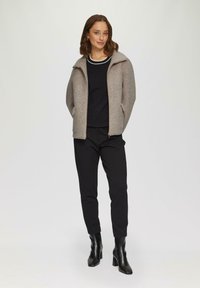 Veste zippée grise avec col haut, portée sur un haut noir et un pantalon noir. Sur fond clair, mettant en valeur les détails de la tenue.