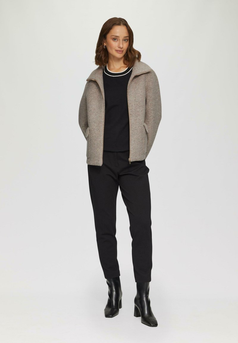 Veste zippée grise avec col haut, portée sur un haut noir et un pantalon noir. Sur fond clair, mettant en valeur les détails de la tenue.