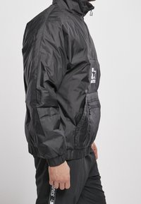 Black windbreaker jacket with a high collar, zippered front, and side pockets. Features elasticized cuffs and a logo detail on the chest.