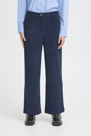Personne portant un pantalon large en velours côtelé bleu marine avec des poches plaquées à l'avant, des bottes noires et une chemise bleu clair boutonnée.
