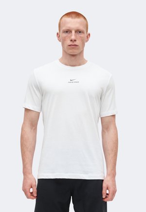 Fiatal férfi rövid vörös hajjal, fehér Nike Pro pólót és fekete rövidnadrágot viselve, karjaival lazán a oldalán áll.